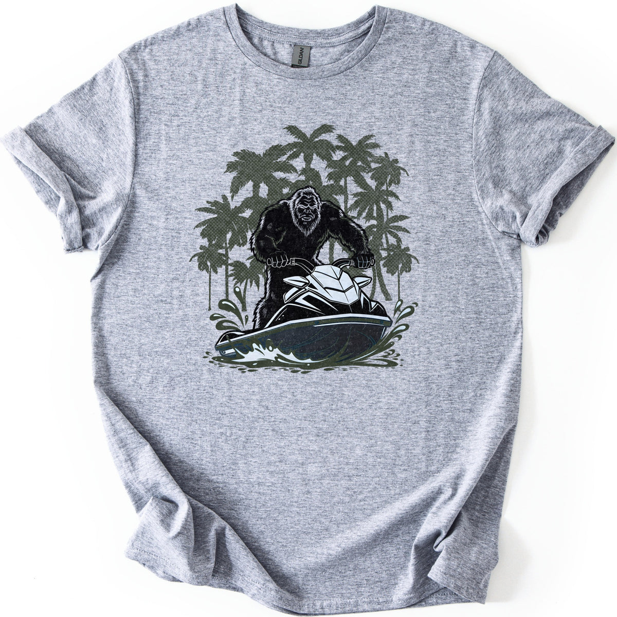 Jet Ski Squatch Comfort Colors T-shirt in Sport Grey, laid flat. A black-furred Bigfoot rides a black and white jet ski through splashing water, with olive green palm trees behind him. No words are printed. The shirt lies on a white background with rolled sleeves.