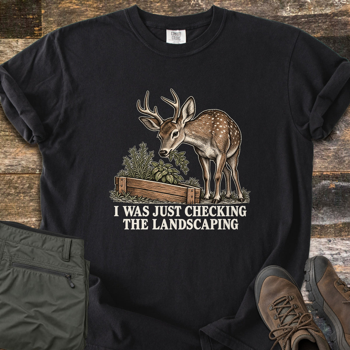 I Was Just Checking the Landscaping Comfort Colors T-shirt in Black, laid flat. A young antlered deer with white spots nibbles herbs from a wooden raised garden bed above the words I Was Just Checking the Landscaping in white. Green pants and brown hiking boots lie beside it on weathered wood.