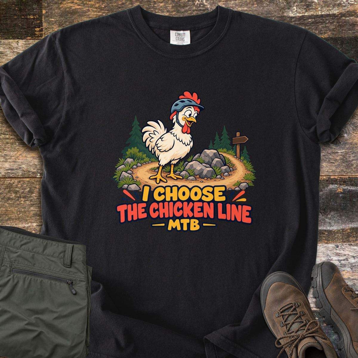 I Choose the Chicken Line MTB Comfort Colors T-shirt in Black, laid flat. A cartoon chicken in a bike helmet stands on a dirt trail by rocks, pine trees and a signpost above the words I Choose the Chicken Line MTB. Olive pants and brown hiking boots lie beside it on wood.
