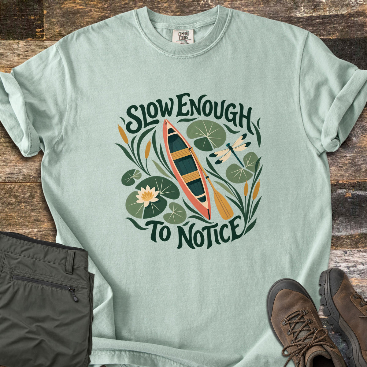 Slow Enough To Notice Comfort Colors T-shirt in Bay, laid flat. A coral canoe with a yellow paddle floats among lily pads, a water lily, cattails and a dragonfly between the words Slow Enough To Notice. Folded green hiking pants and brown hiking boots sit on weathered wood.