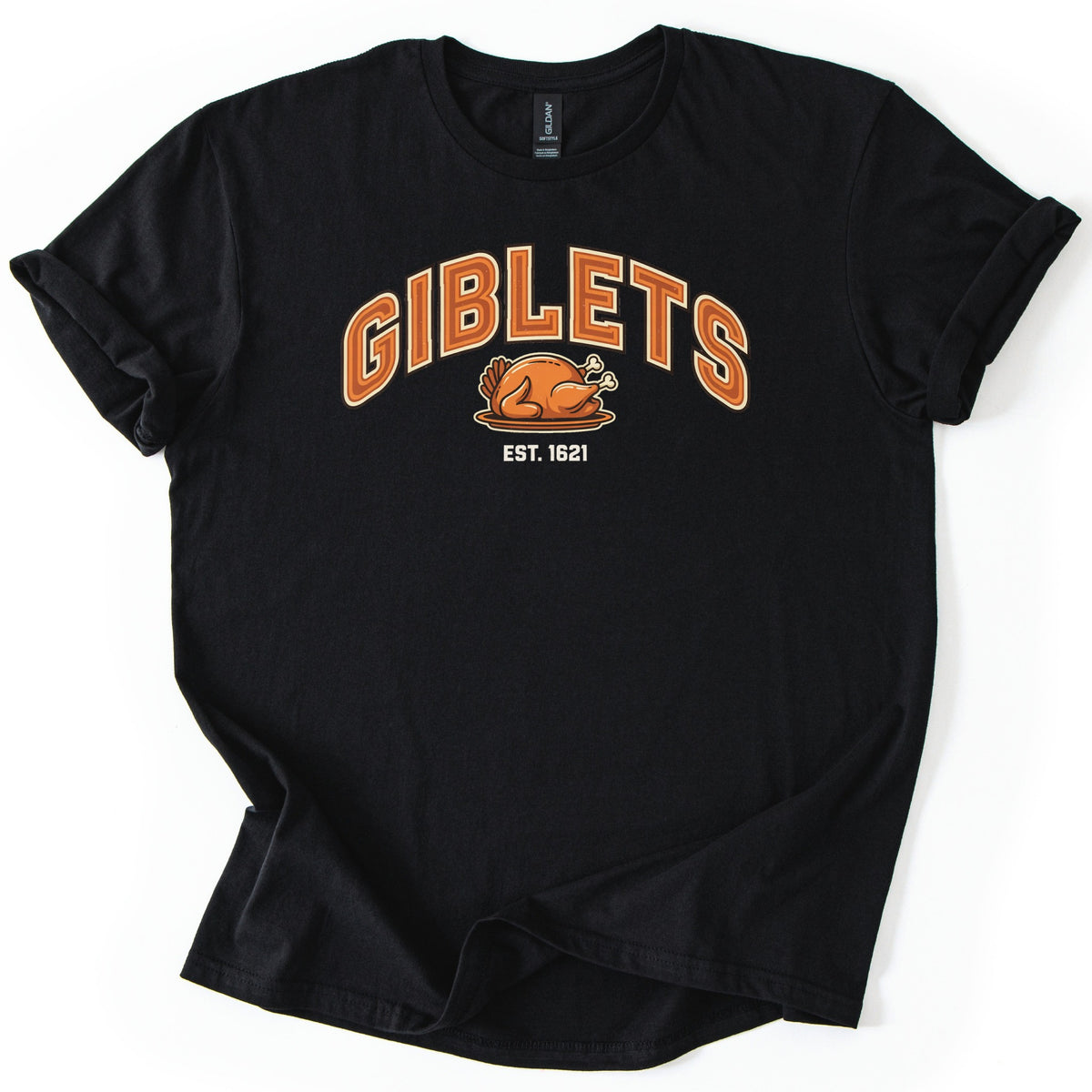 Giblets Comfort Colors T-shirt in Black, laid flat. Arched orange varsity-style letters read GIBLETS above a cartoon roast turkey on a platter, with EST. 1621 in white below. The shirt has rolled sleeves and lies on a white background.