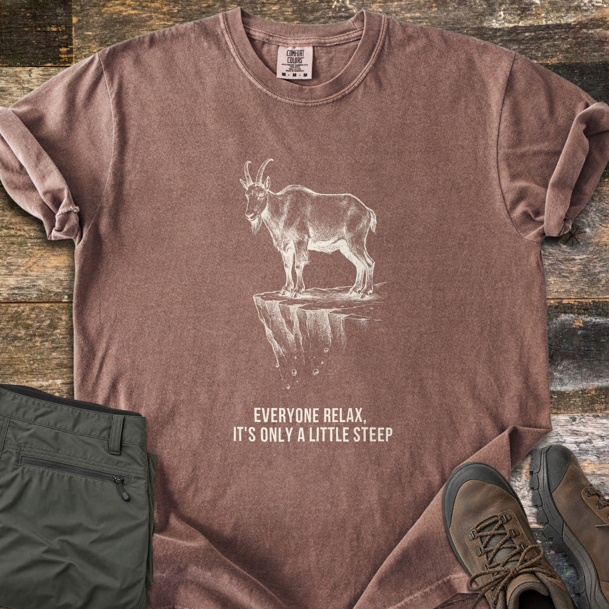 Everyone Relax It's Only A Little Steep Comfort Colors T-shirt in Espresso, laid flat. A white sketch of a horned mountain goat standing on a crumbling cliff ledge above the words Everyone Relax, It's Only A Little Steep. Sleeves are rolled, on wood planks with green hiking pants and brown boots.