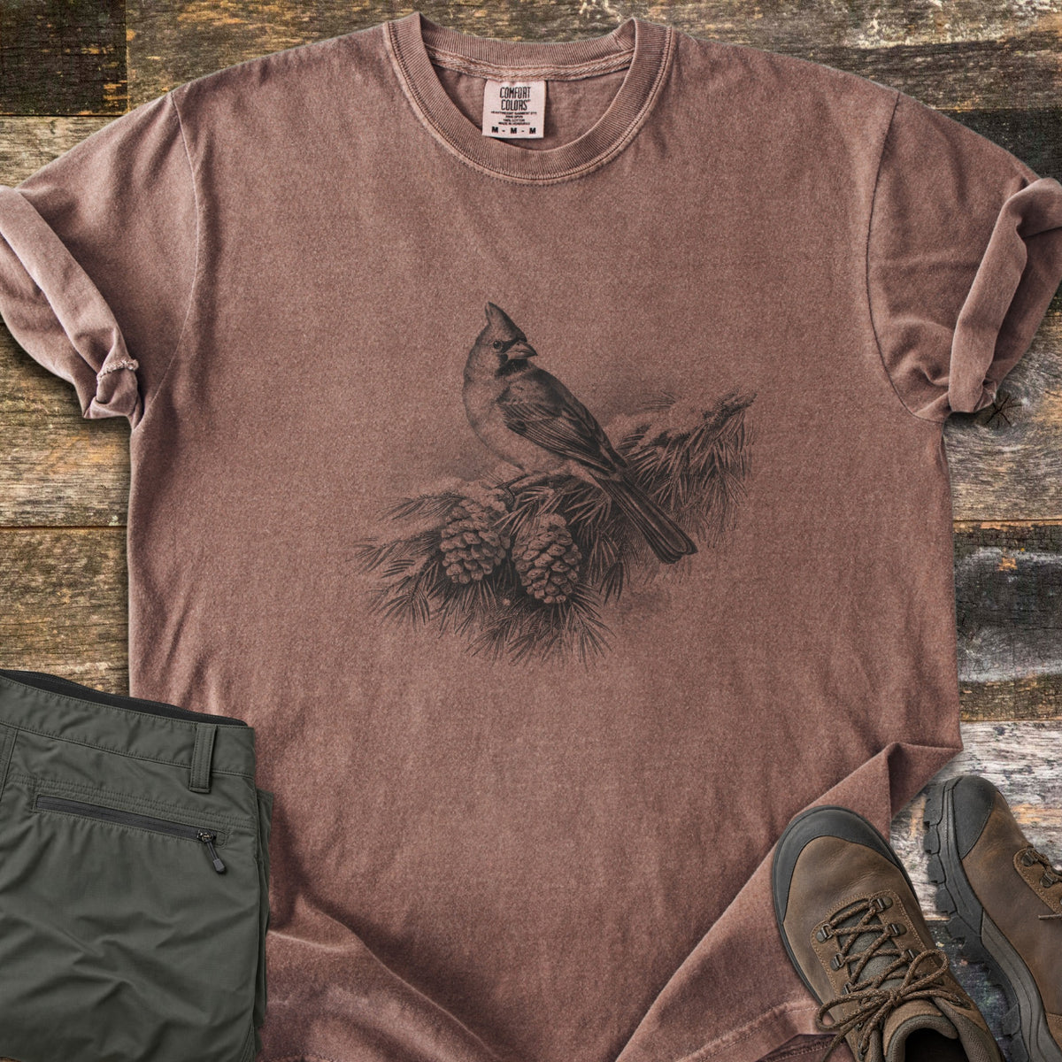 Cardinal On A Snowy Bough Comfort Colors T-shirt in Espresso, laid flat. A grayscale drawing of a cardinal perched on a snow-covered pine branch with two pinecones, with no words. Sleeves rolled, on weathered wood planks beside olive hiking pants and brown hiking boots.