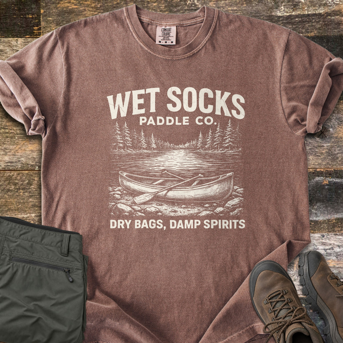 Wet Socks Paddle Co Comfort Colors T-shirt in Espresso, laid flat. Cream print reads Wet Socks Paddle Co. above a sketched canoe and paddle on a rocky lakeshore with pines, then Dry Bags, Damp Spirits. Olive hiking pants and brown boots sit on weathered wood.