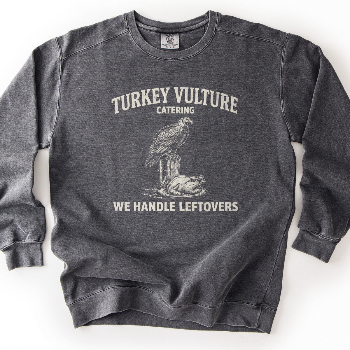 Turkey Vulture Catering Comfort Colors crewneck sweatshirt in Pepper, laid flat. Cream print of a vulture perched on a wooden post above a roast turkey on a platter, with the words Turkey Vulture Catering above and We Handle Leftovers below. Shown on a white background.