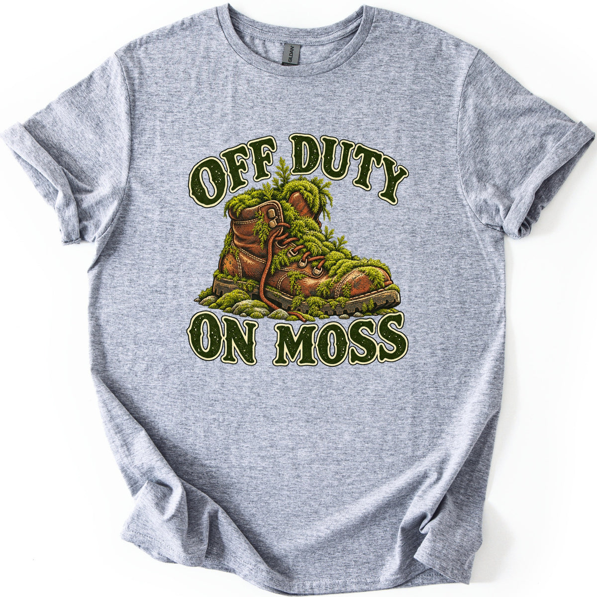 Off Duty on Moss Comfort Colors T-shirt in Sport Grey, laid flat. A worn brown leather hiking boot overgrown with green moss and ferns sits on mossy stones, with OFF DUTY arched above and ON MOSS below in dark green letters. The shirt lies on a white surface with cuffed sleeves.