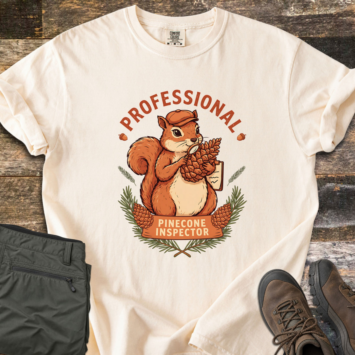 Professional Pinecone Inspector Comfort Colors T-shirt in Ivory, laid flat. A cartoon squirrel in an orange cap studies a pinecone with a magnifying glass, holding a clipboard, under the word Professional, with Pinecone Inspector on a banner. Hiking pants and boots lie on wood.