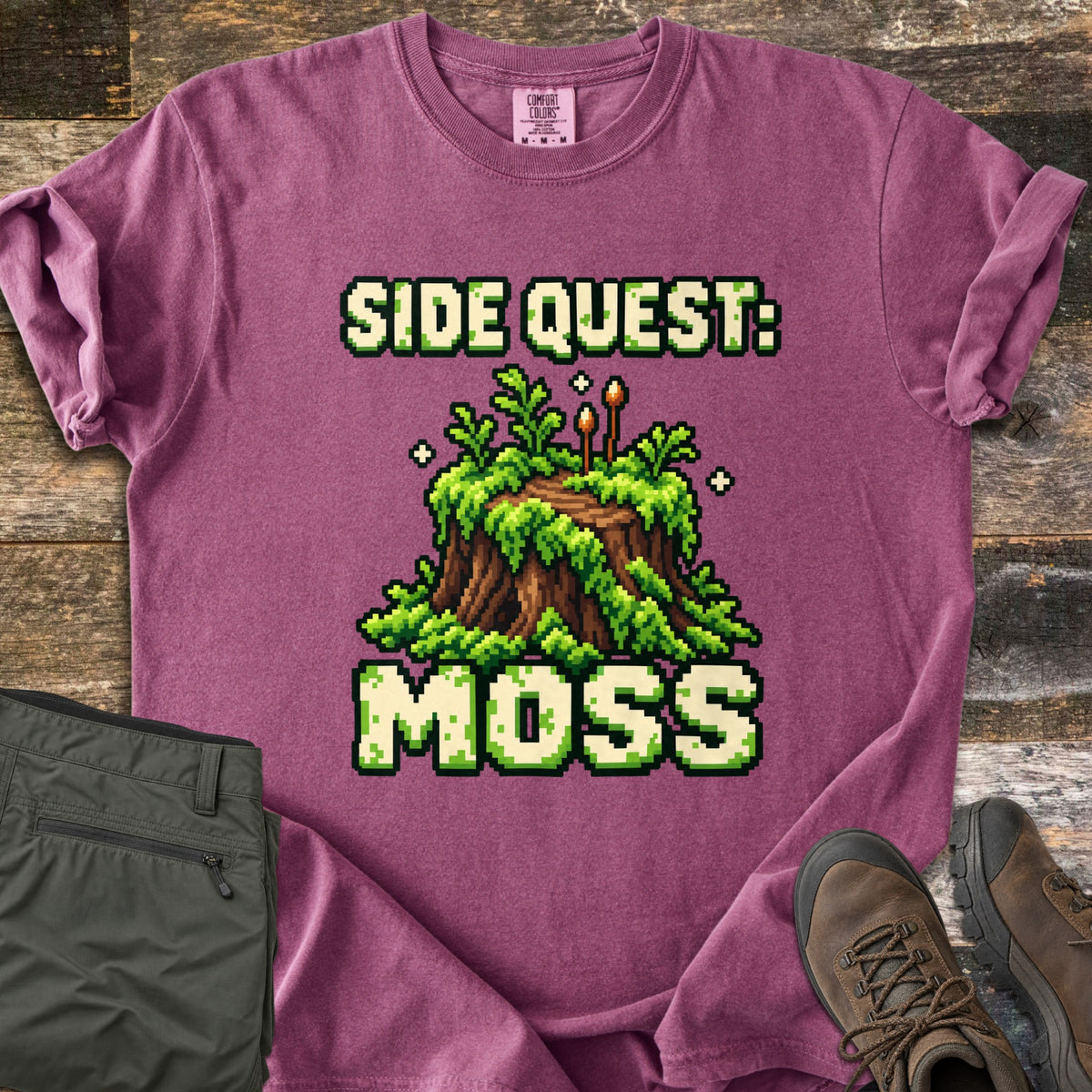 Side Quest Moss Comfort Colors T-shirt in Berry, laid flat. Pixel art of a moss-covered tree stump with ferns, two small sporophytes and sparkles, between the words Side Quest: and Moss. Olive cargo pants and brown hiking boots lie beside it on weathered wood planks.