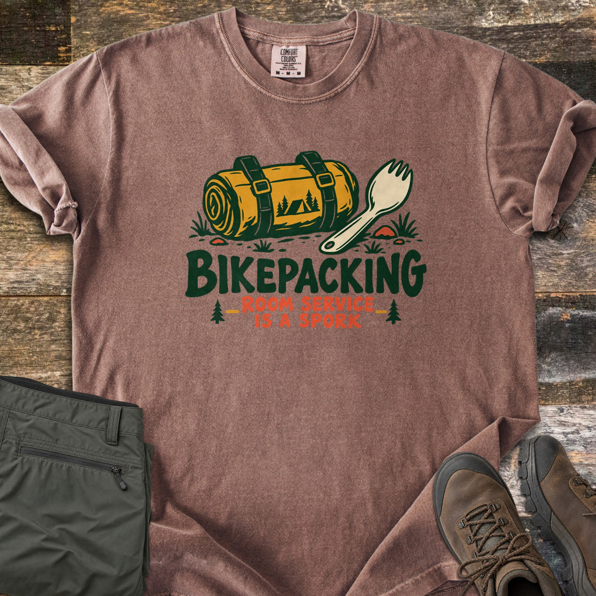 Bikepacking Room Service Is a Spork Comfort Colors T-shirt in Espresso, laid flat. A yellow bedroll with dark green straps and a cream spork sit on grass above the words Bikepacking Room Service Is a Spork. Folded olive pants and brown hiking boots lie beside it on weathered wood.