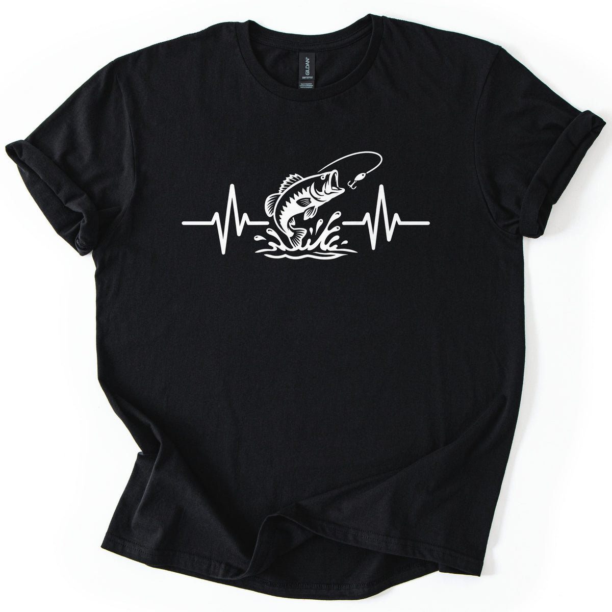 Fishing Heartbeat Comfort Colors T-shirt in Black, laid flat. A white line drawing of a bass jumping out of splashing water at a lure on a fishing line breaks up a heartbeat line across the chest. No words are printed. The shirt lies on a white surface with cuffed sleeves.