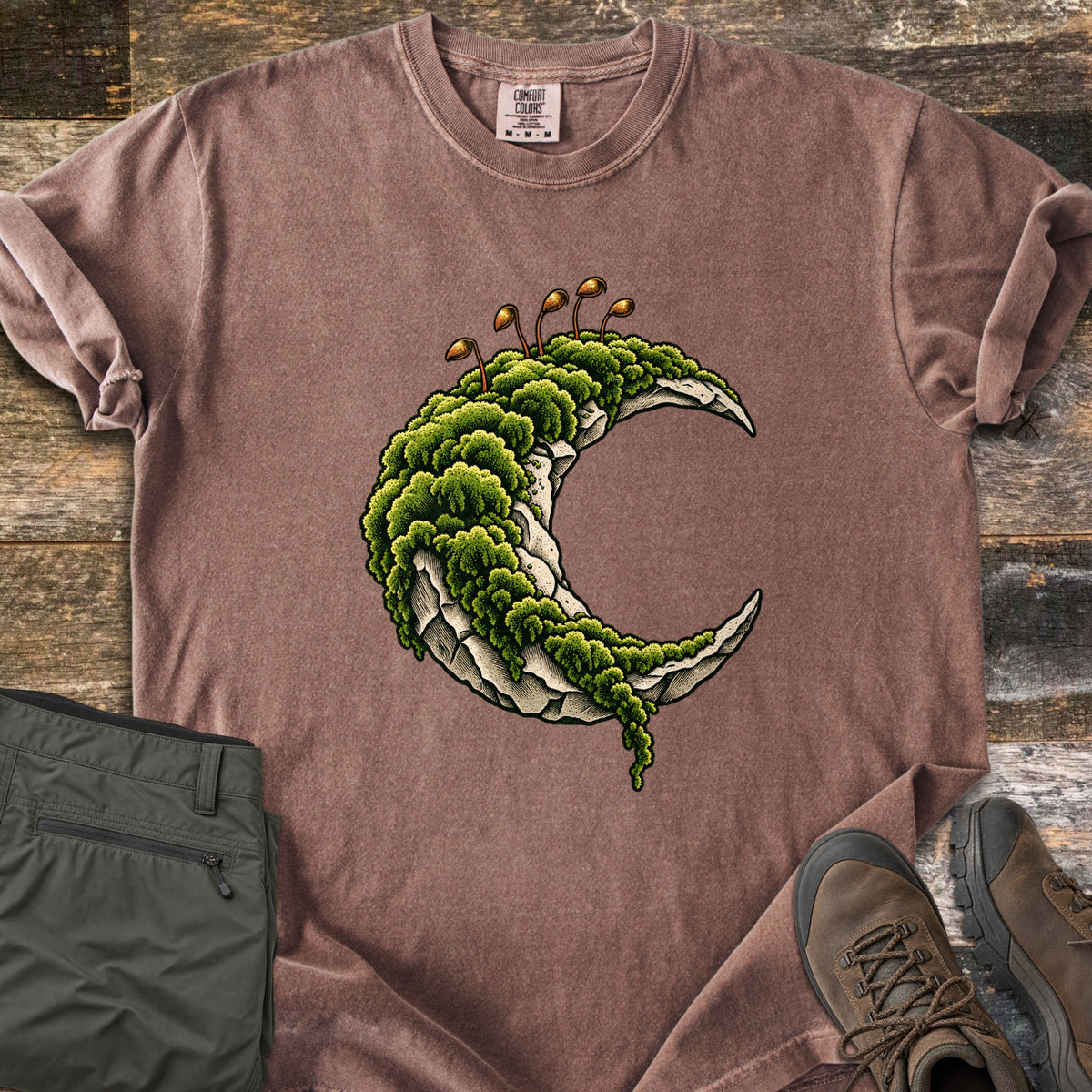 Moss Moon Comfort Colors T-shirt in Espresso, laid flat. A pale stone crescent moon covered in thick green moss, with thin brown spore stalks rising from the top. Sleeves rolled, on weathered wood planks beside folded olive hiking pants and brown hiking boots.
