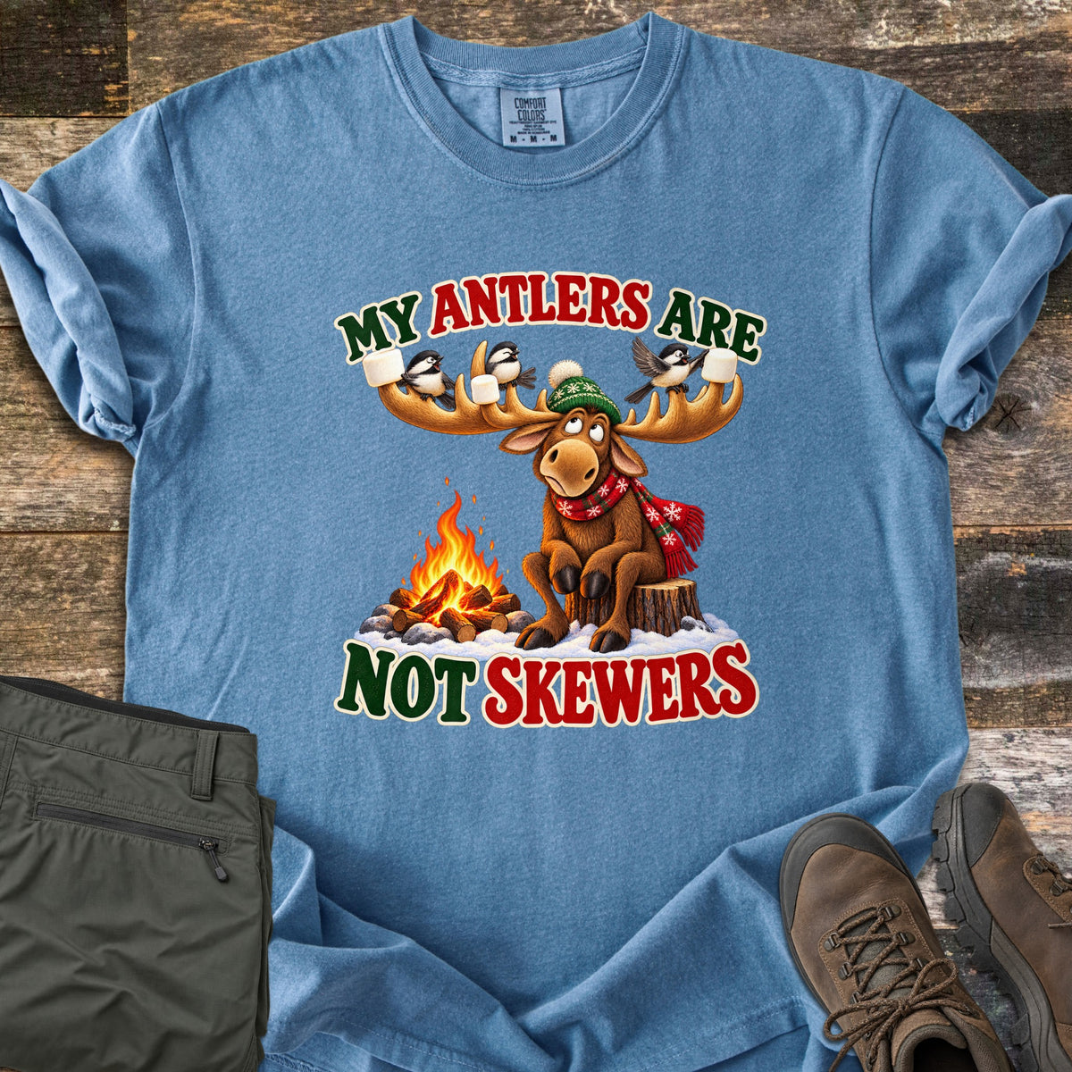 My Antlers Are Not Skewers Comfort Colors T-shirt in Blue Jean, laid flat. A cartoon moose in a green knit hat and red scarf sits on a stump by a campfire as chickadees on its antlers hold marshmallows, with the words My Antlers Are Not Skewers. Olive pants and brown boots lie on weathered wood.