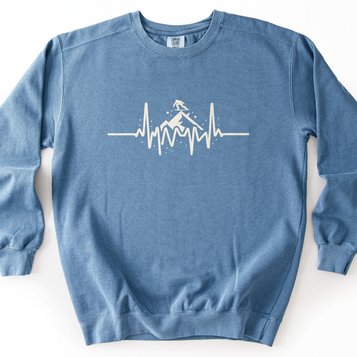 Snow Boarder Heartbeat Comfort Colors crewneck sweatshirt in Blue Jean, laid flat. A cream heartbeat line runs across the chest, rising into a mountain peak with a snowboarder riding on top and small dots of snow around it. No words. The sweatshirt lies on a white surface.