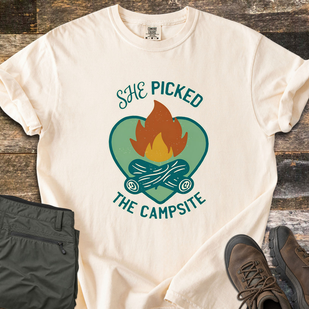 She Picked The Campsite Comfort Colors T-shirt in Ivory, laid flat. A campfire with crossed logs sits on a green heart, with the words She Picked above and The Campsite below in teal. Sleeves are rolled on a rustic wood floor beside folded olive hiking pants and brown hiking boots.