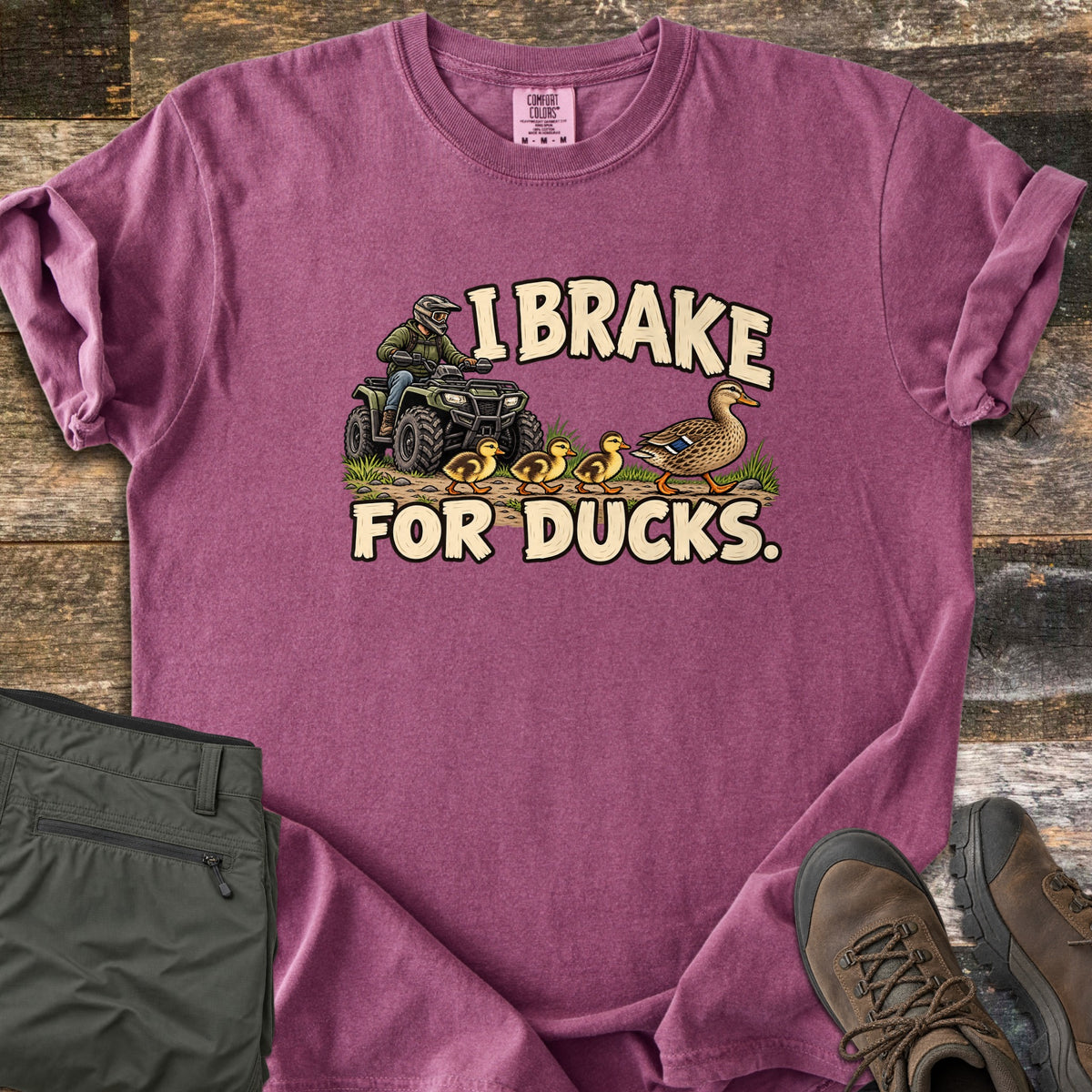 I Brake for Ducks Comfort Colors T-shirt in Berry, laid flat. A rider in a helmet on a green ATV waits as a mother duck leads her ducklings across a dirt path, under the words I Brake For Ducks. Olive pants and brown hiking boots lie beside it on weathered wood planks.
