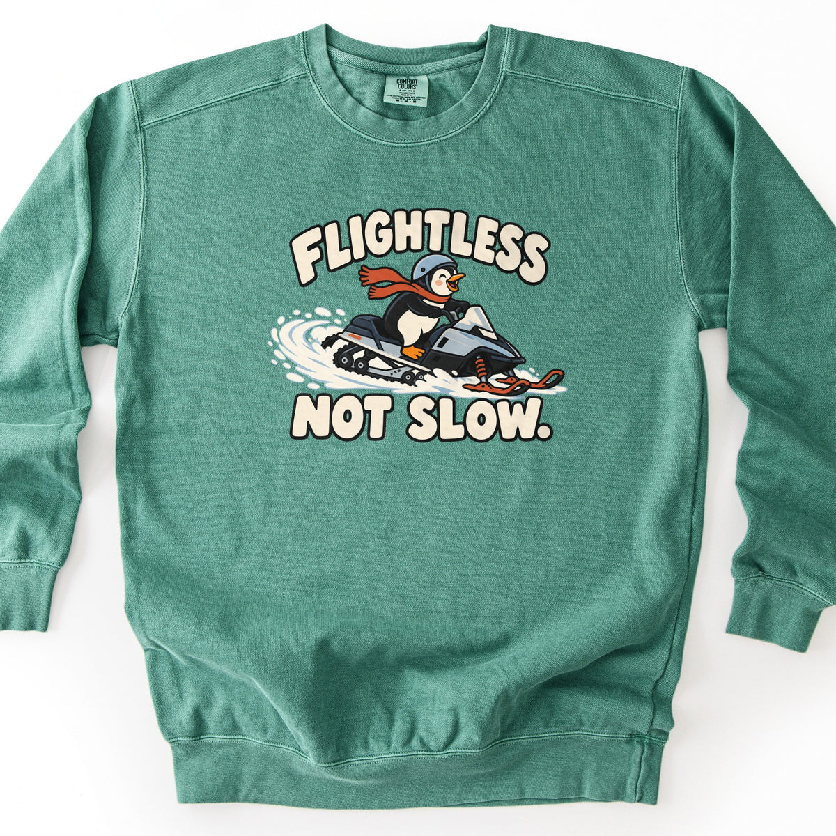 Flightless Not Slow Comfort Colors crewneck sweatshirt in Light Green, laid flat. A cartoon penguin wearing a helmet and red scarf speeds on a snowmobile with orange skis through spraying snow, between the cream words Flightless and Not Slow. It lies on a white background.