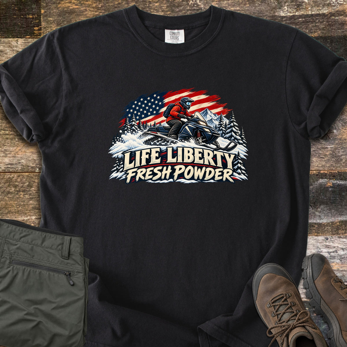 Life Liberty Fresh Powder Comfort Colors T-shirt in Black, laid flat. A rider in a red jacket and helmet drives a snowmobile through snowy pines past mountains and a waving American flag, above the words Life Liberty Fresh Powder. Gray pants and brown hiking boots lie on weathered wood.