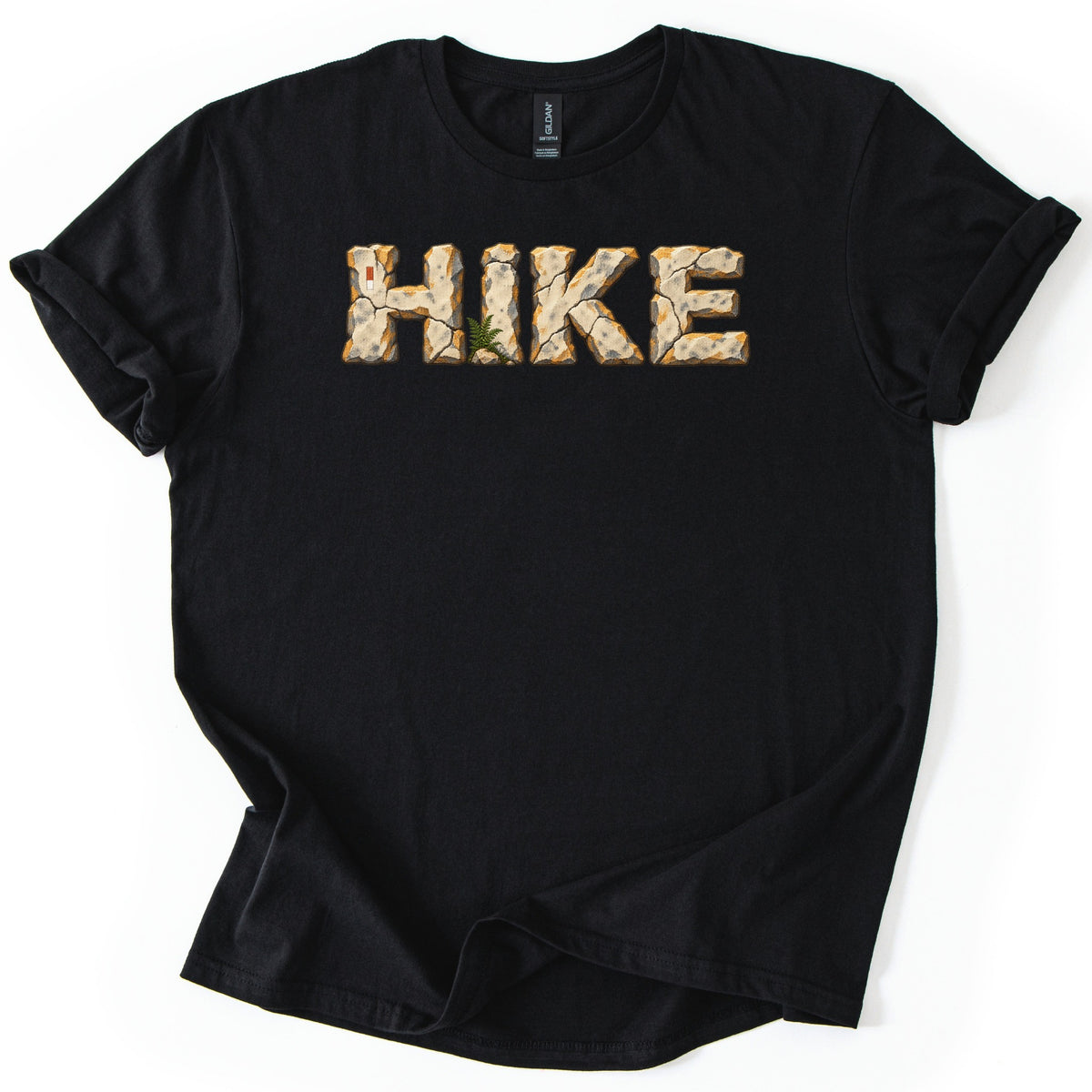 Hike Comfort Colors T-shirt in Black, laid flat. The word HIKE is printed in letters made of cracked tan stone with orange edges, a small green fern at the base and a red and white trail marker on the H. Sleeves rolled, on a white background.
