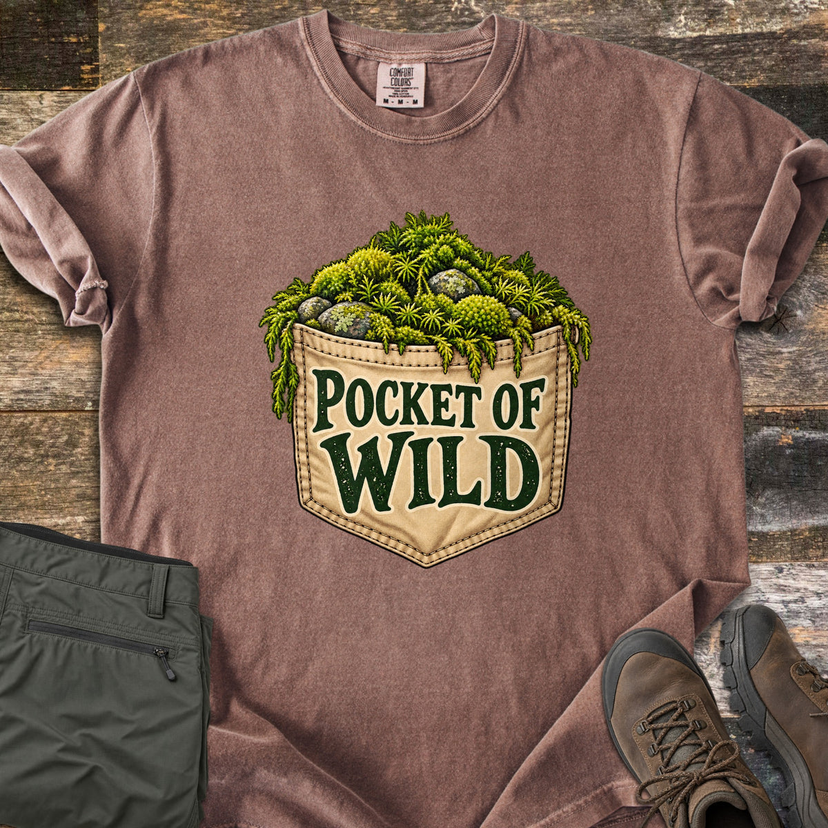 Pocket of Wild Comfort Colors T-shirt in Espresso, laid flat. A tan stitched jeans pocket overflowing with green moss and lichen-covered stones, printed with the words Pocket of Wild. Shown on weathered wood with folded olive hiking pants and brown hiking boots.