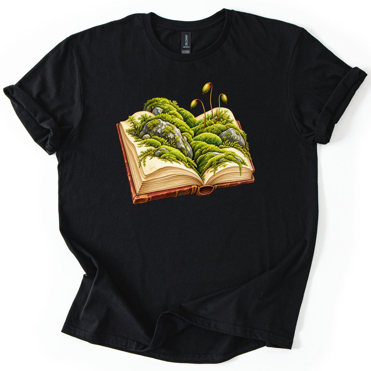 Moss Storybook Comfort Colors T-shirt in Black, laid flat. An open old book with a worn red-brown cover holds mounds of green moss and gray rocks on its pages, with three thin spore stalks on top. The shirt lies on a white background with its sleeves cuffed.