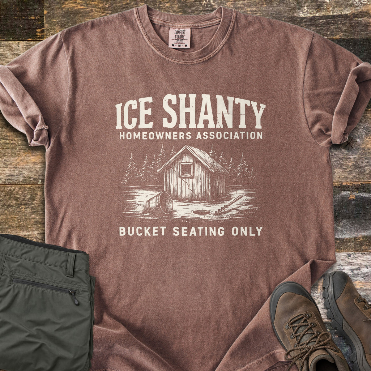 Ice Shanty Homeowners Association Comfort Colors T-shirt in Espresso, laid flat. Cream print of a wooden ice shanty among pines, a tipped bucket, ice hole and auger, with the words Ice Shanty Homeowners Association and Bucket Seating Only. Olive pants and brown hiking boots sit on weathered wood.