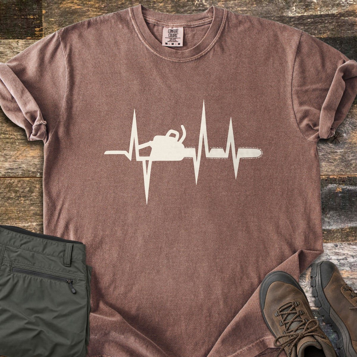 Chainsaw Heartbeat Comfort Colors T-shirt in Espresso, laid flat. A cream heartbeat line runs across the chest with a chainsaw silhouette built into it, the bar and chain extending to the right. Folded olive cargo shorts and brown hiking boots sit on a weathered wood floor.