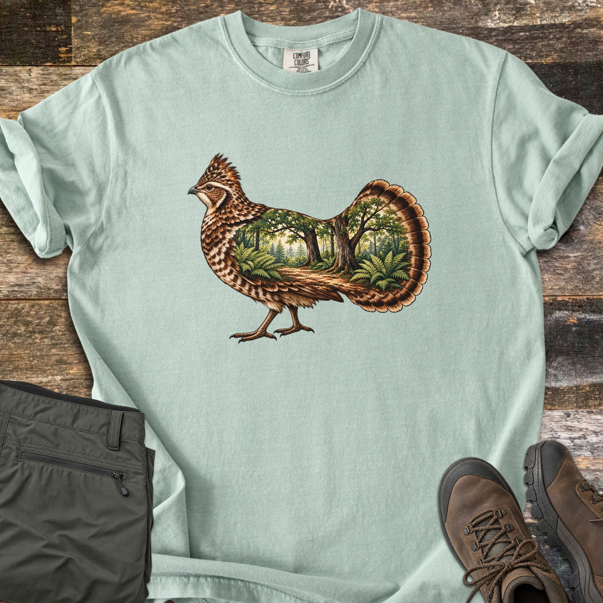 Ruffed Grouse Woods Comfort Colors T-shirt in Bay, laid flat. A detailed ruffed grouse with a fanned tail, its body filled with a forest scene of oak trees, ferns and a dirt path. Folded olive pants and brown hiking boots sit beside it on weathered wood planks.