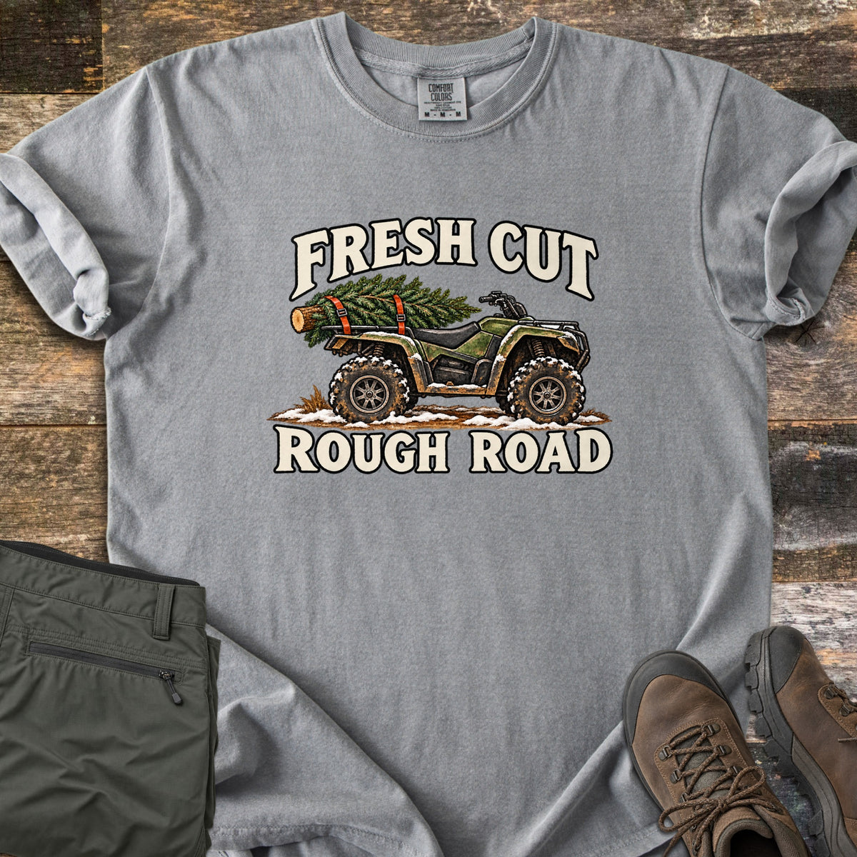 Fresh Cut Rough Road Comfort Colors T-shirt in Grey, laid flat. An army-green ATV with muddy tires carries a strapped-on pine tree over snowy ground, between the words Fresh Cut and Rough Road. Shown on wood planks with olive cargo pants and brown hiking boots.