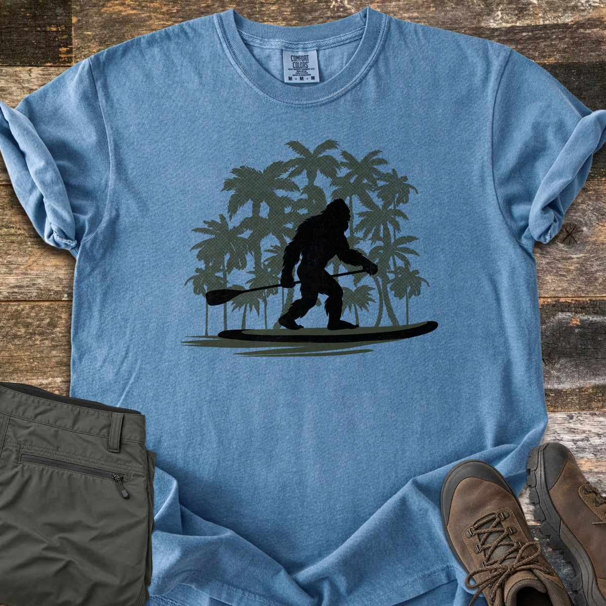 Paddle Board Squatch Comfort Colors T-shirt in Blue Jean, laid flat. A black silhouette of Bigfoot standing on a paddle board and holding a paddle, in front of dark green palm trees. Olive pants and brown hiking boots lie beside the shirt on weathered wood planks.