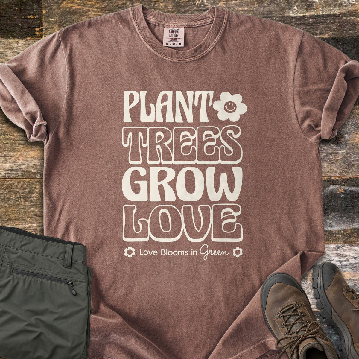 Plant Trees Grow Love Comfort Colors T-shirt in Espresso, laid flat. Cream retro bubble letters read Plant Trees Grow Love beside a smiling daisy, above Love Blooms in Green. Sleeves cuffed on weathered wood with folded olive hiking pants and brown hiking boots.