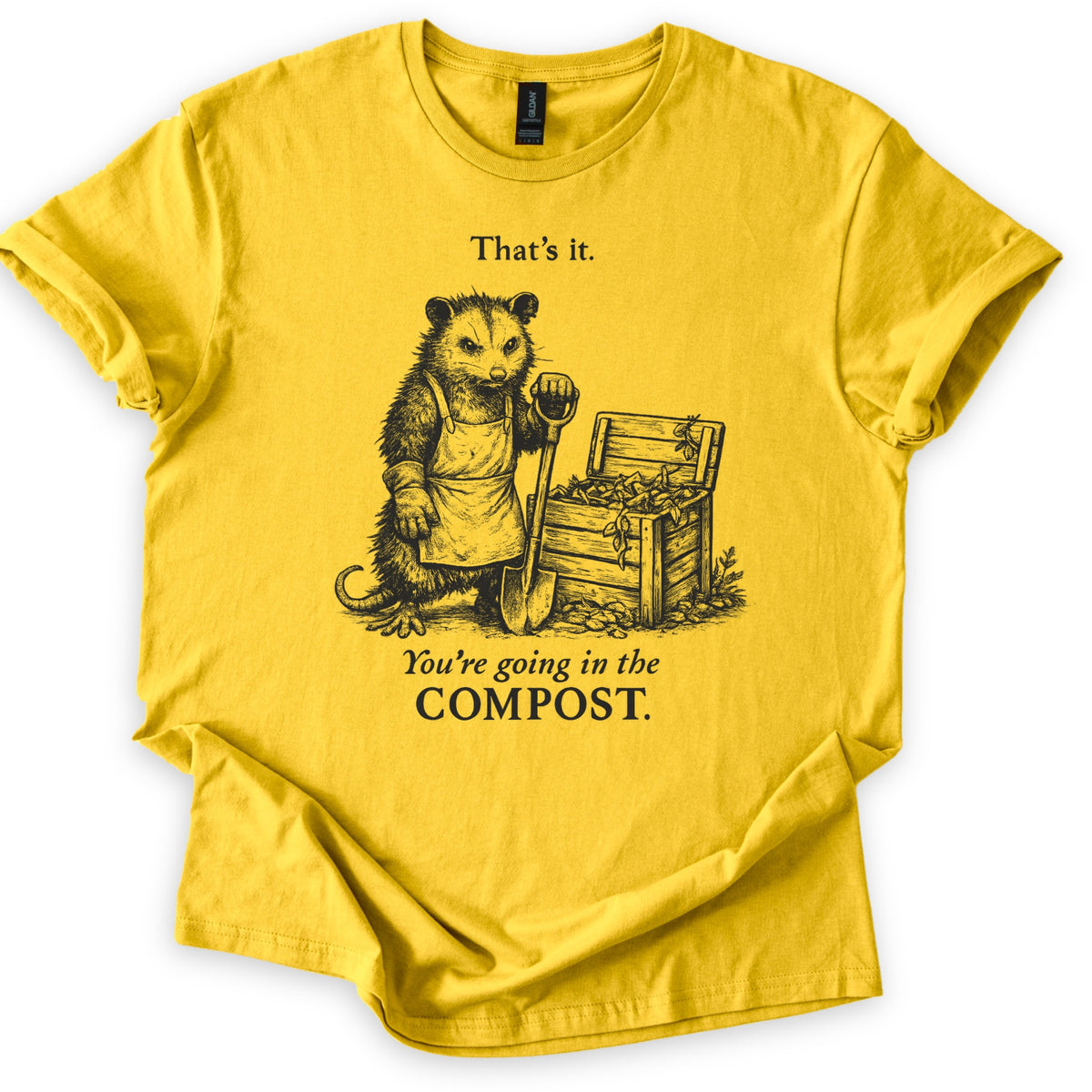 Going In The Compost Possum Comfort Colors T-shirt in Daisy, laid flat. A scowling possum in an apron and work glove leans on a shovel beside an open wooden compost bin, between the words That's it. and You're going in the COMPOST. Black print, shirt on white with cuffed sleeves.