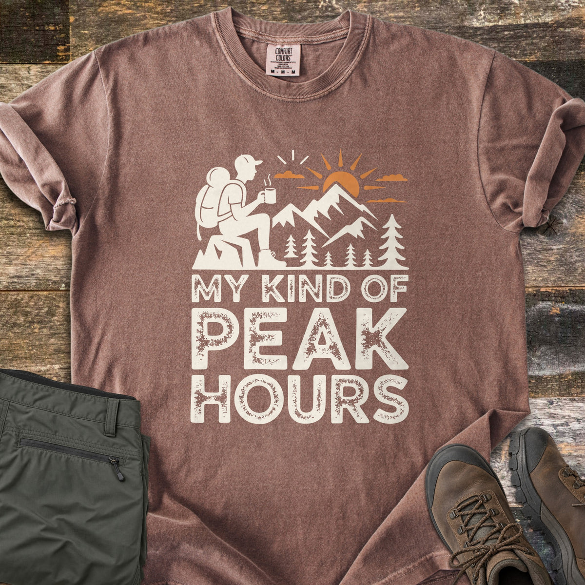 My Kind Of Peak Hours Comfort Colors T-shirt in Espresso, laid flat. A cream hiker with a backpack sits on a rock with a steaming mug, facing mountains, pines and an orange sun, above the words My Kind Of Peak Hours. Olive hiking pants and brown boots lie on weathered wood.