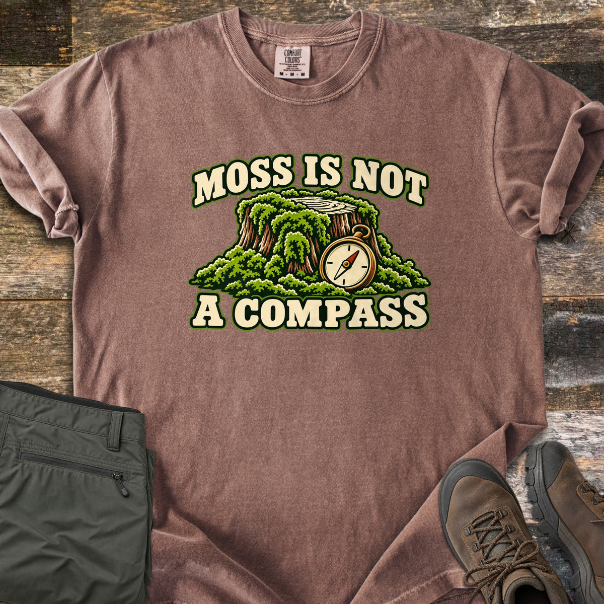 Moss Is Not a Compass Comfort Colors T-shirt in Espresso, laid flat. A moss-covered tree stump with a brass compass leaning on it sits between the words MOSS IS NOT and A COMPASS. Olive hiking pants and brown hiking boots lie on rustic wood planks.