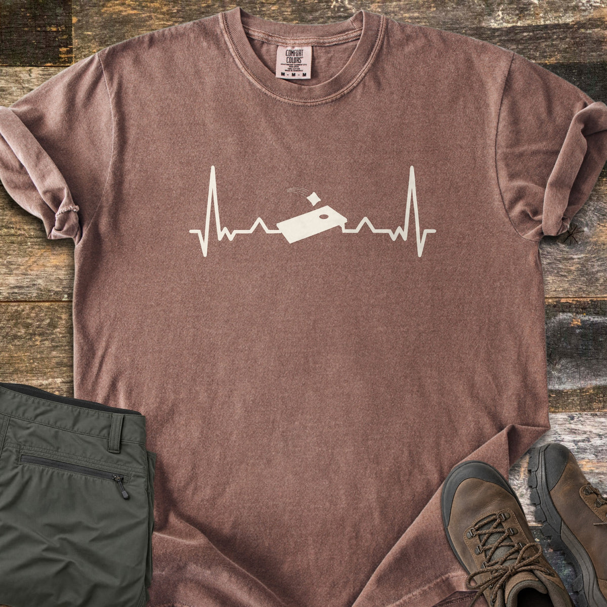 Corn Hole Comfort Colors T-shirt in Espresso, laid flat. A cream heartbeat line runs across the chest with a cornhole board at its center and a bean bag flying toward the hole. Olive pants and brown hiking boots sit beside it on weathered wood planks.