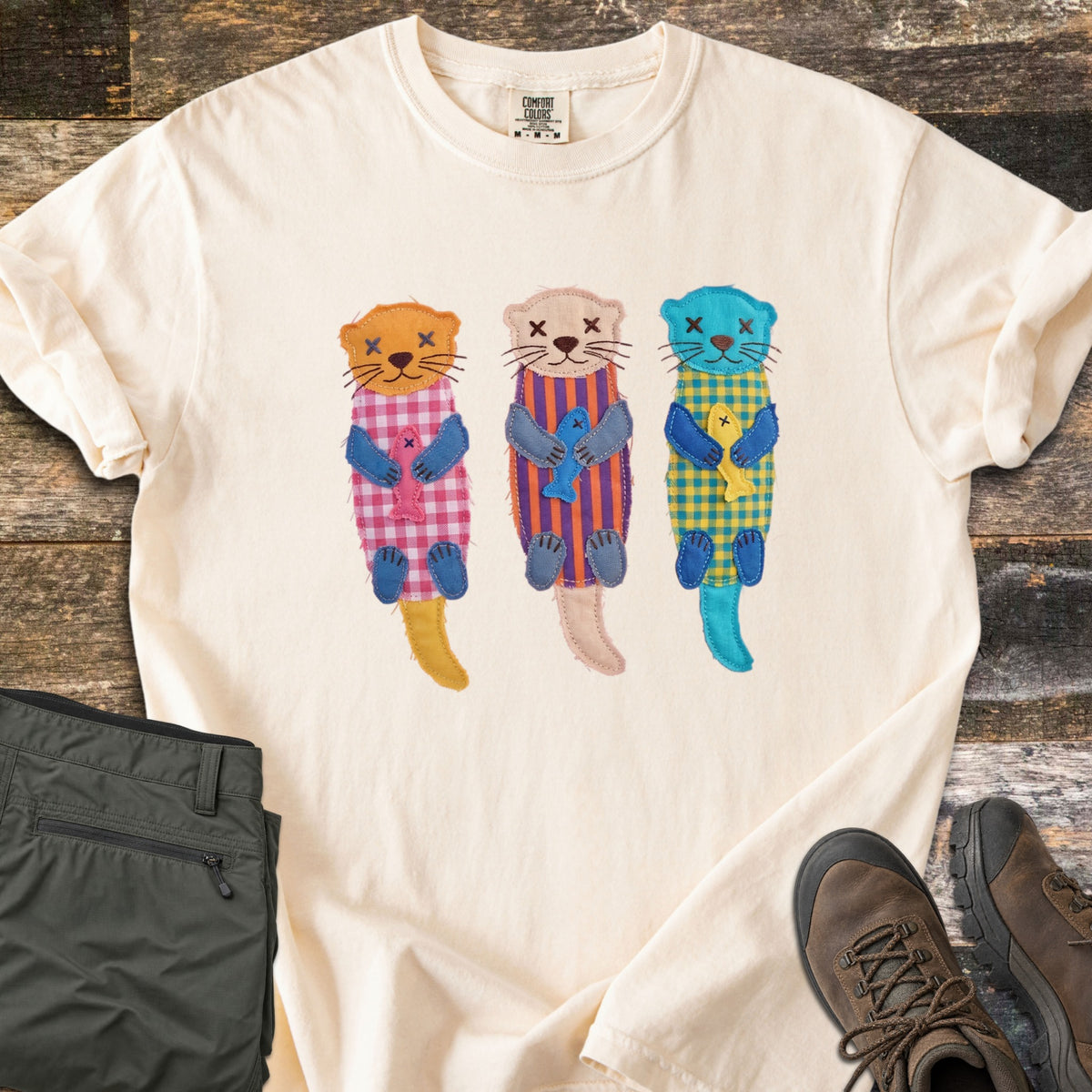Patchwork Otter Comfort Colors T-shirt in Ivory, laid flat. Three patchwork otters in orange, peach and turquoise float on their backs, each in a gingham or striped body hugging a small fish, with X-stitched eyes. Olive pants and brown hiking boots sit on a wood floor.
