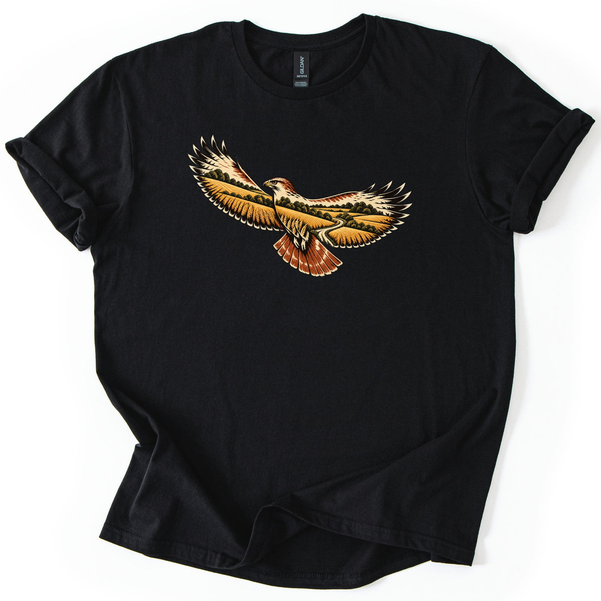 Hawk Over Golden Fields Comfort Colors T-shirt in Black, laid flat. A hawk in flight with spread wings showing a landscape of golden fields, rolling hills, trees and a winding path inside its feathers, and a rust-red tail. No words. Sleeves rolled, on a white surface.