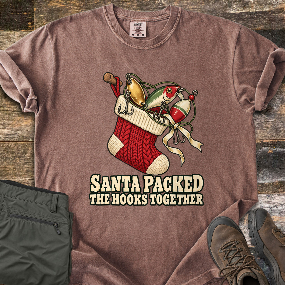 Santa Packed The Hooks Together Comfort Colors T-shirt in Espresso, laid flat. A red knit Christmas stocking holds fishing lures, a bobber and a rod handle tied with a cream ribbon above the words Santa Packed The Hooks Together. Olive pants and brown boots sit on a wood floor.