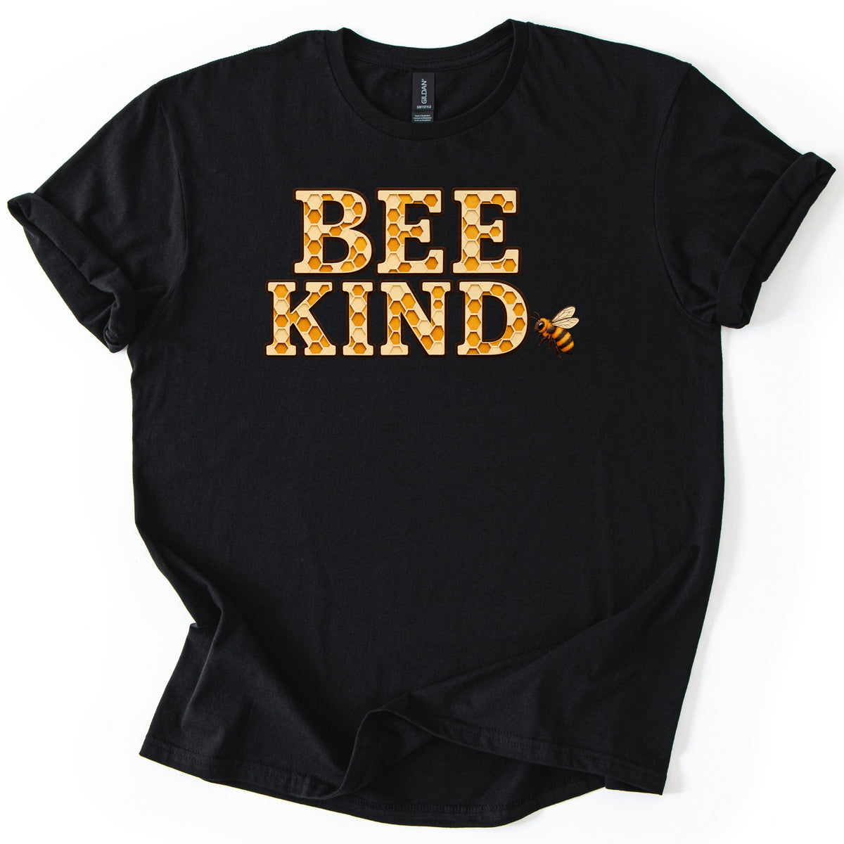 Bee Kind Comfort Colors T-shirt in Black, laid flat. The words BEE KIND in large block letters filled with a golden honeycomb pattern and outlined in brown, with a small honeybee beside the D. The shirt has rolled sleeves and lies on a white background.