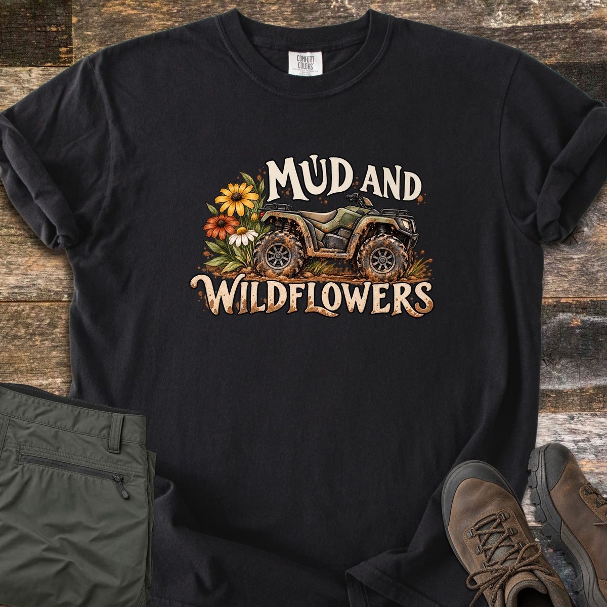 Mud and Wildflowers Comfort Colors T-shirt in Black, laid flat. A mud-splattered green ATV beside yellow, orange and white wildflowers, with the words Mud and Wildflowers above and below. Folded olive pants and brown hiking boots sit on a weathered wood background.