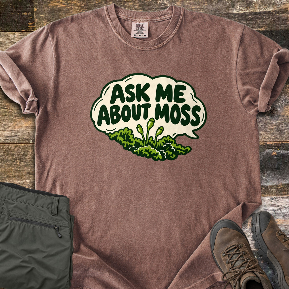 Ask Me About Moss Comfort Colors T-shirt in Espresso, laid flat. A cream speech bubble reads Ask Me About Moss in dark green letters above a mound of green moss with three spore stalks. Sleeves rolled, on weathered wood with folded olive hiking pants and brown hiking boots.