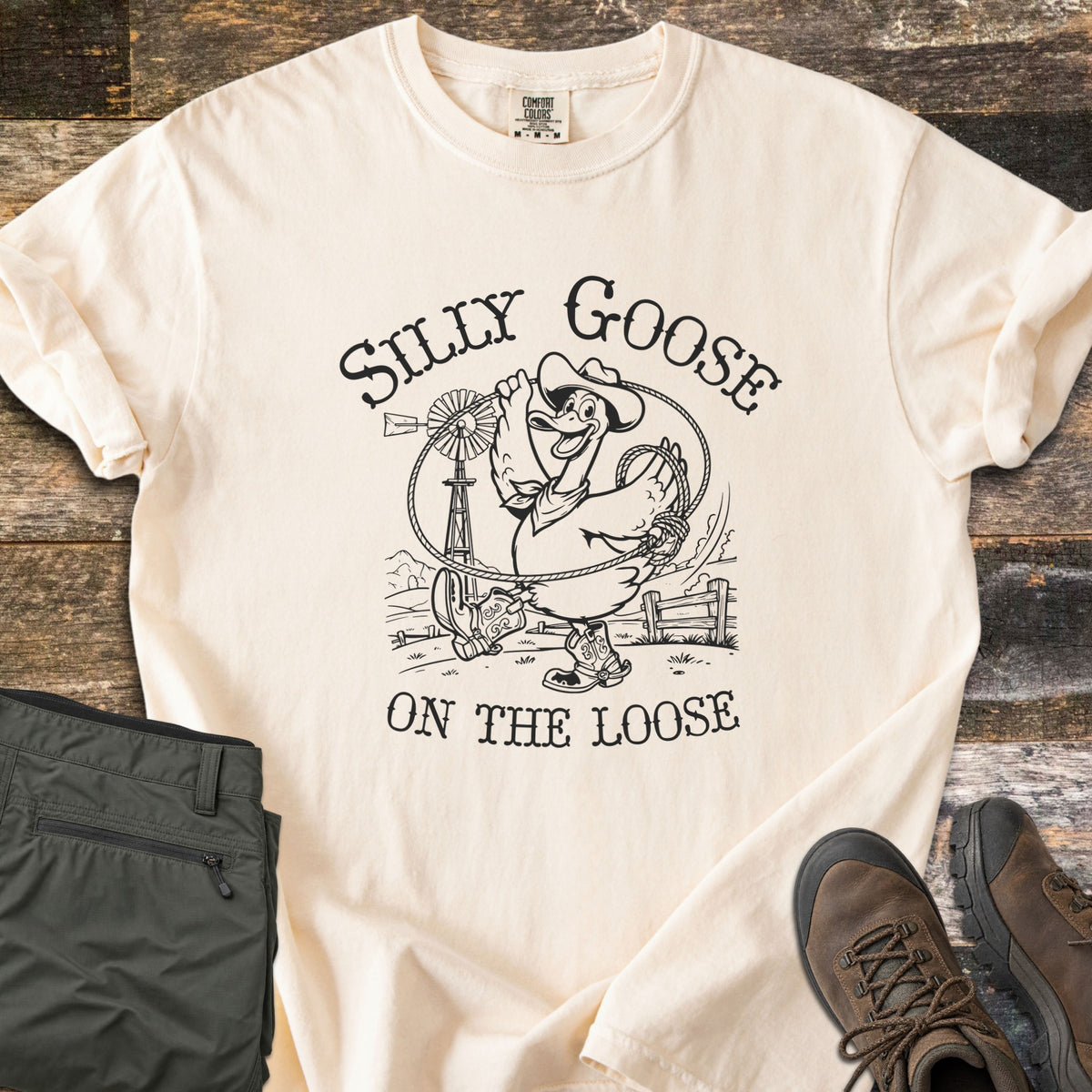Silly Goose on the Loose Comfort Colors T-shirt in Ivory, laid flat. Black line art of a grinning cartoon goose in a cowboy hat, bandana, and boots swinging a lasso by a windmill and fence, between the words Silly Goose and On the Loose. Olive pants and brown boots sit on wood planks.