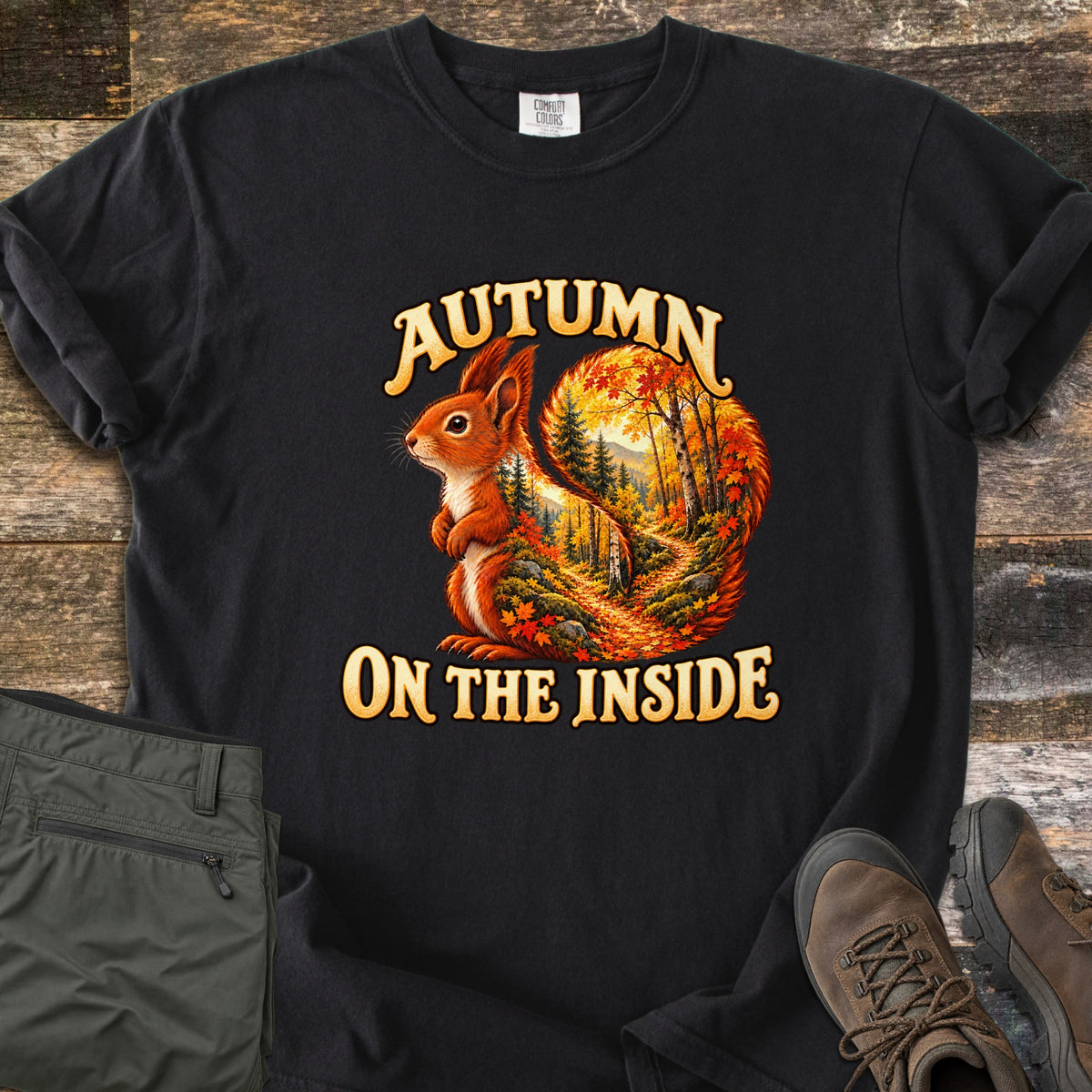 Autumn on the Inside Comfort Colors T-shirt in Black, laid flat. A red squirrel whose tail frames a forest path through orange and yellow autumn trees, with the words Autumn on the Inside. Olive cargo pants and brown hiking boots lie beside it on weathered wood.
