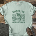 Sounds Like Someone Else's Problem Comfort Colors T-shirt in Bay, laid flat. Green sketch-style art of a capybara sitting in a pond with eyes closed, under the words Sounds Like and above Someone Else's Problem. Olive pants and brown hiking boots lie on a dark wood surface.