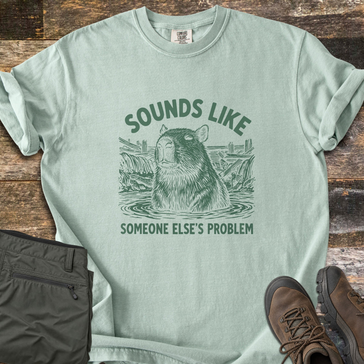 Sounds Like Someone Else's Problem Comfort Colors T-shirt in Bay, laid flat. Green sketch-style art of a capybara sitting in a pond with eyes closed, under the words Sounds Like and above Someone Else's Problem. Olive pants and brown hiking boots lie on a dark wood surface.