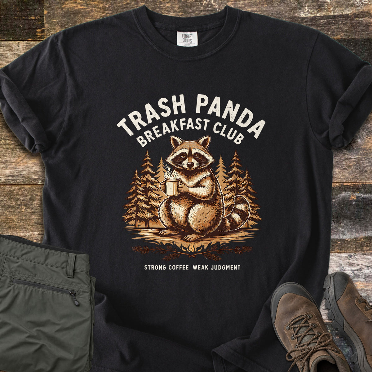 Trash Panda Breakfast Club Comfort Colors T-shirt in Black, laid flat. A raccoon holding a steaming mug sits among pine trees under the words Trash Panda Breakfast Club in cream, with Strong Coffee Weak Judgment below. Folded green pants and brown hiking boots lie on weathered wood.