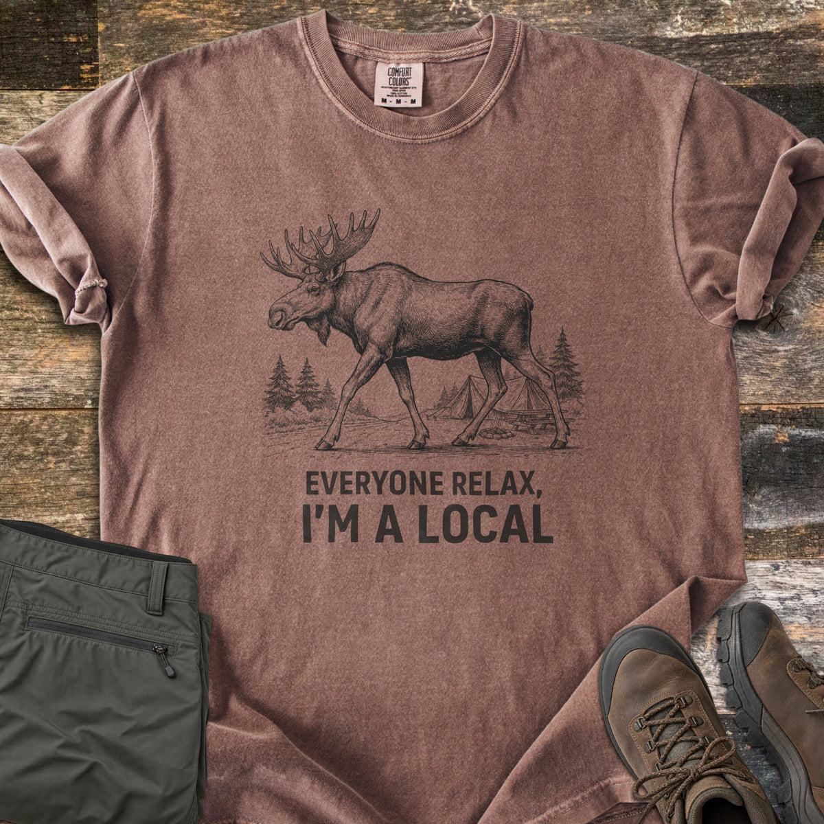 Everyone Relax I'm A Local Comfort Colors T-shirt in Espresso, laid flat. A sketched moose walks past pine trees and tents at a campsite above the words Everyone Relax, I'm A Local. Sleeves are rolled on a wood floor beside folded green pants and brown hiking boots.