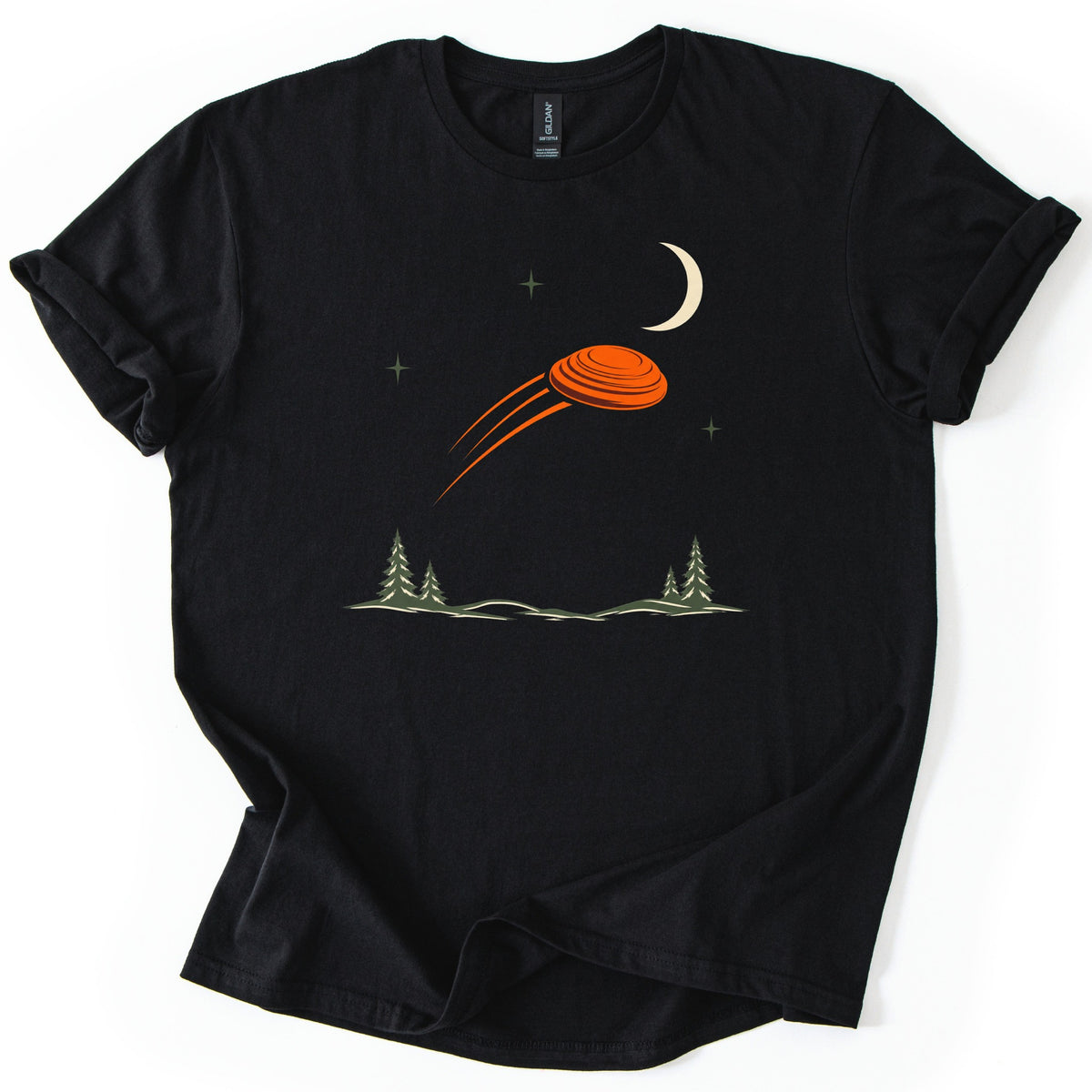 Moonrise Clay Comfort Colors T-shirt in Black, laid flat. An orange clay target streaks across a night sky with a cream crescent moon and small green stars, above pine trees and low hills. No words are printed. Sleeves rolled, on a white background.