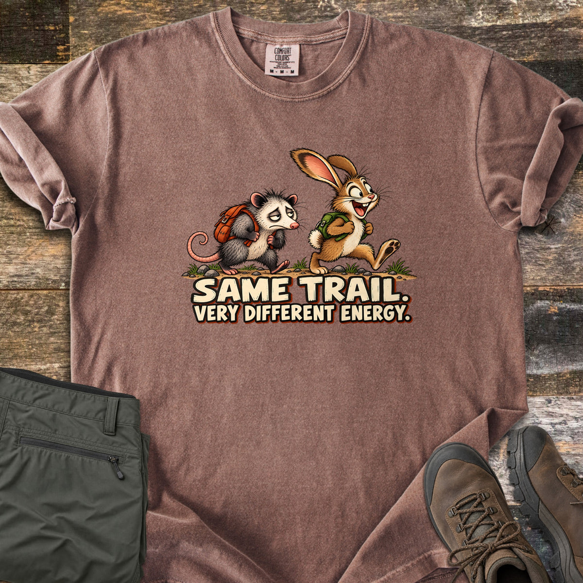 Same Trail. Very Different Energy. Comfort Colors T-shirt in Espresso, laid flat. A tired cartoon opossum with an orange backpack trudges behind a grinning, bounding rabbit with a green backpack on a dirt trail, above the words Same Trail. Very Different Energy. Shown on wood with green pants and hiking boots.