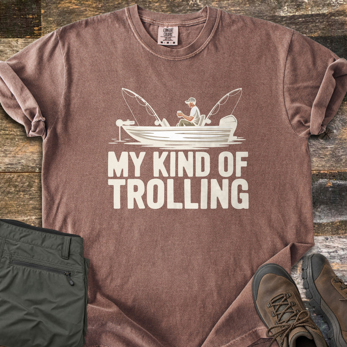 My Kind Of Trolling Comfort Colors T-shirt in Espresso, laid flat. A man in a cap holds a cup in a small fishing boat with two bent rods and motors, above the words My Kind Of Trolling in cream. Olive cargo pants and brown hiking boots sit beside it on weathered wood.