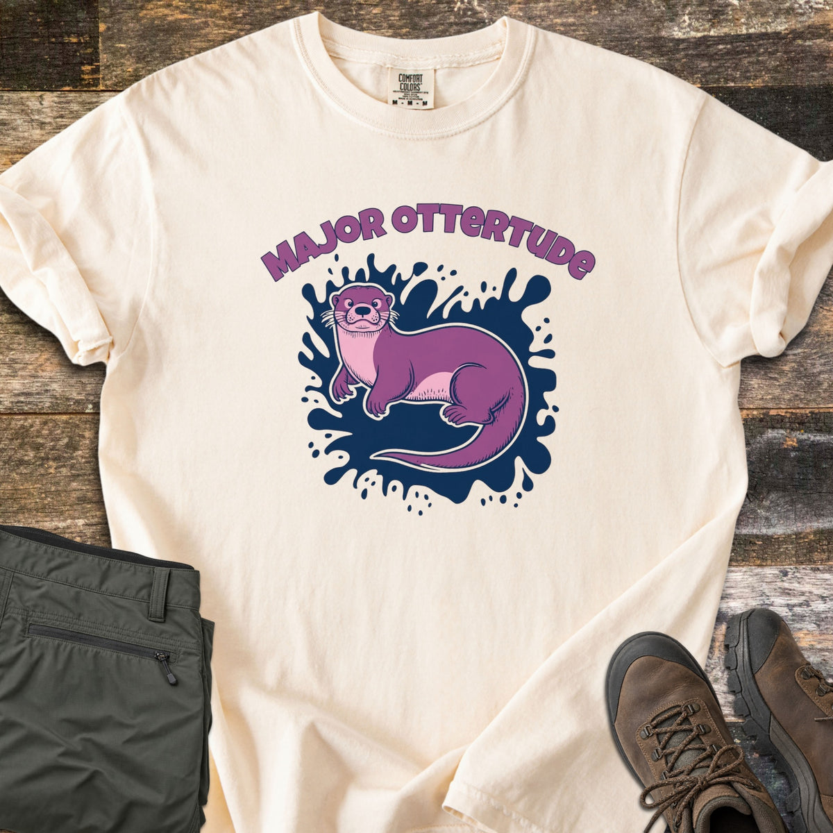 Major Ottertude Comfort Colors T-shirt in Ivory, laid flat. A purple cartoon otter lies on a navy paint splash beneath the arched words Major Ottertude. Sleeves are rolled, on weathered wood with folded olive pants and brown hiking boots.